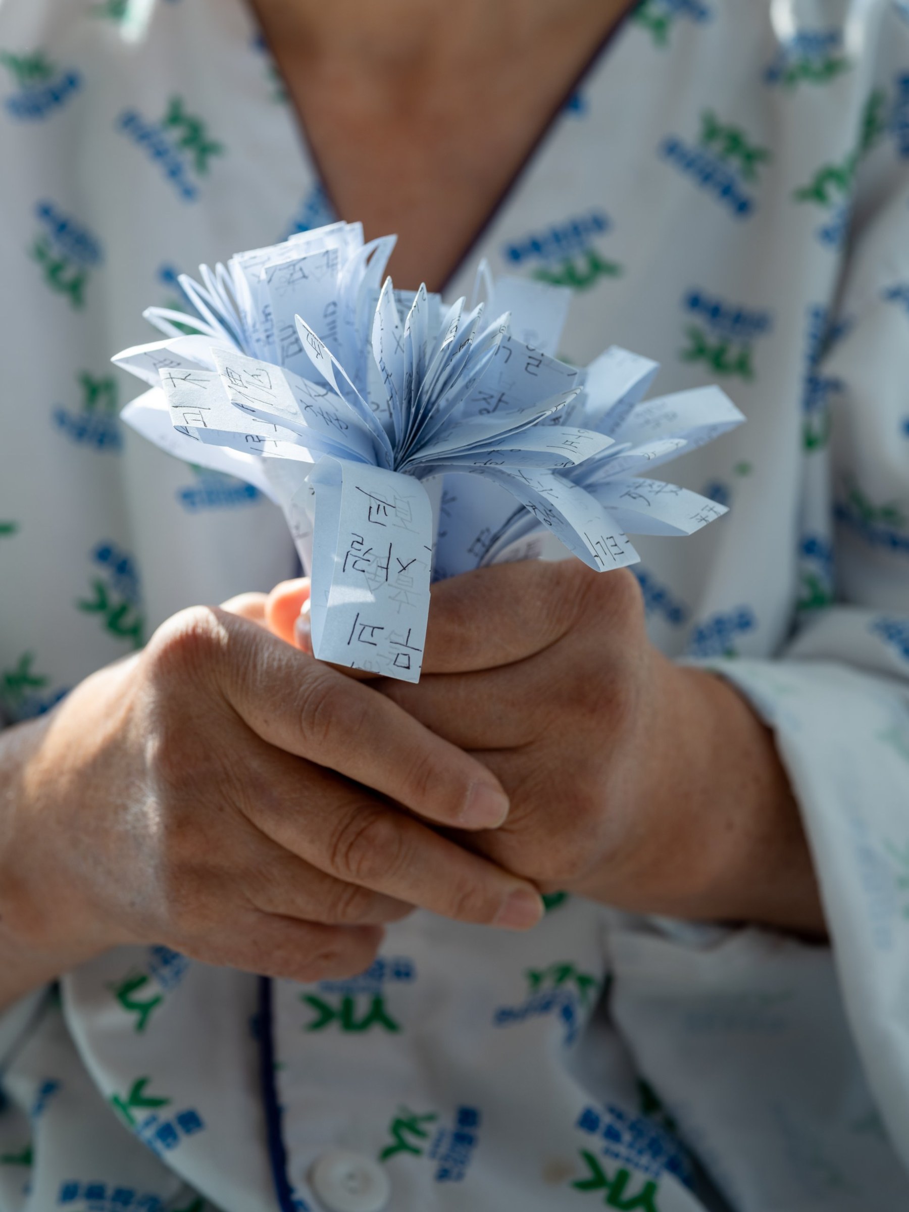 병원복을 입은 사람이 손글씨가 적힌 종이로 접은 꽃을 두 손으로 쥐고 있다