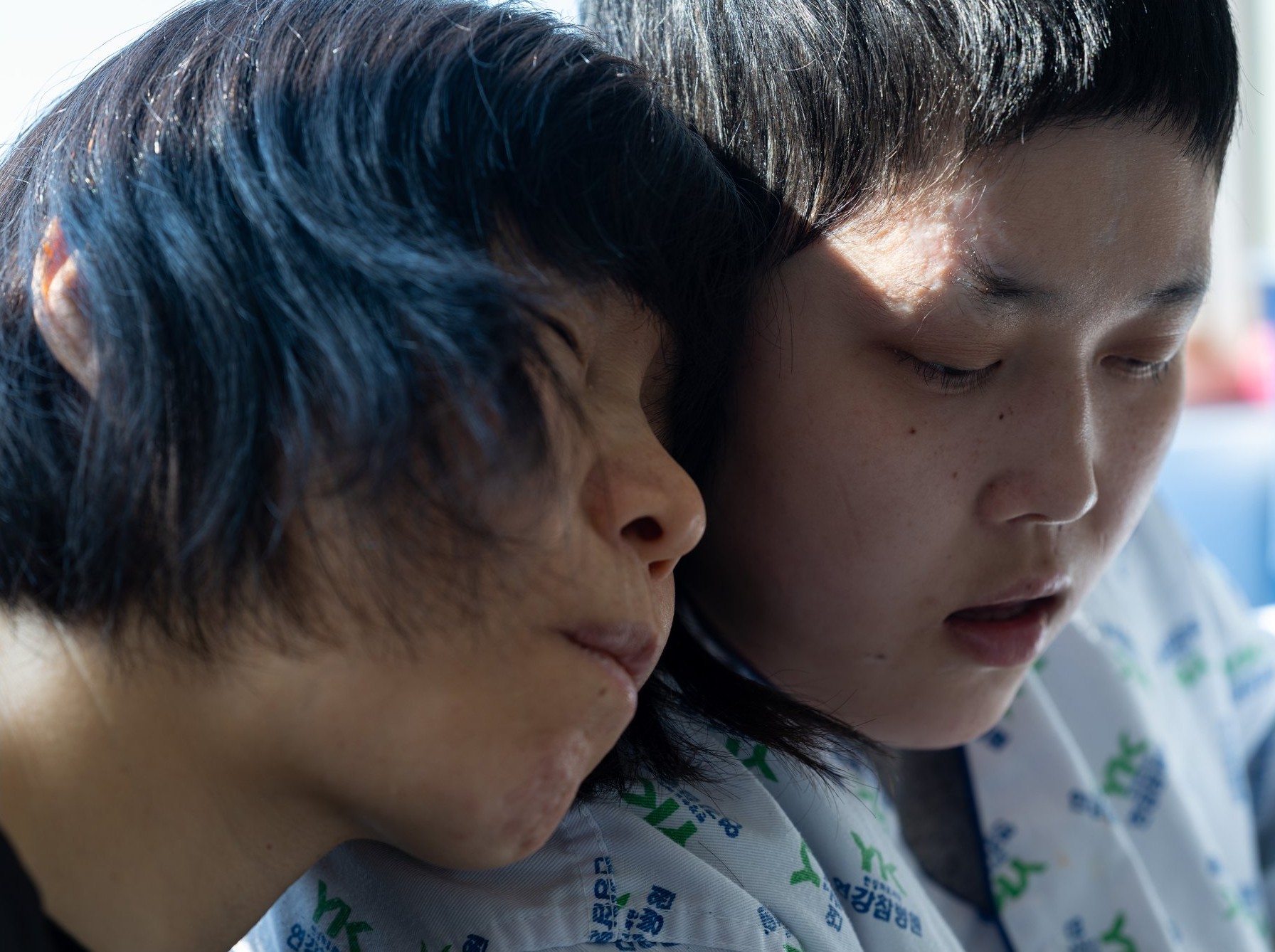 병원복을 입은 두 사람, 한 사람이 눈을 감고 다른 사람의 머리에 얼굴을 기대고 있다